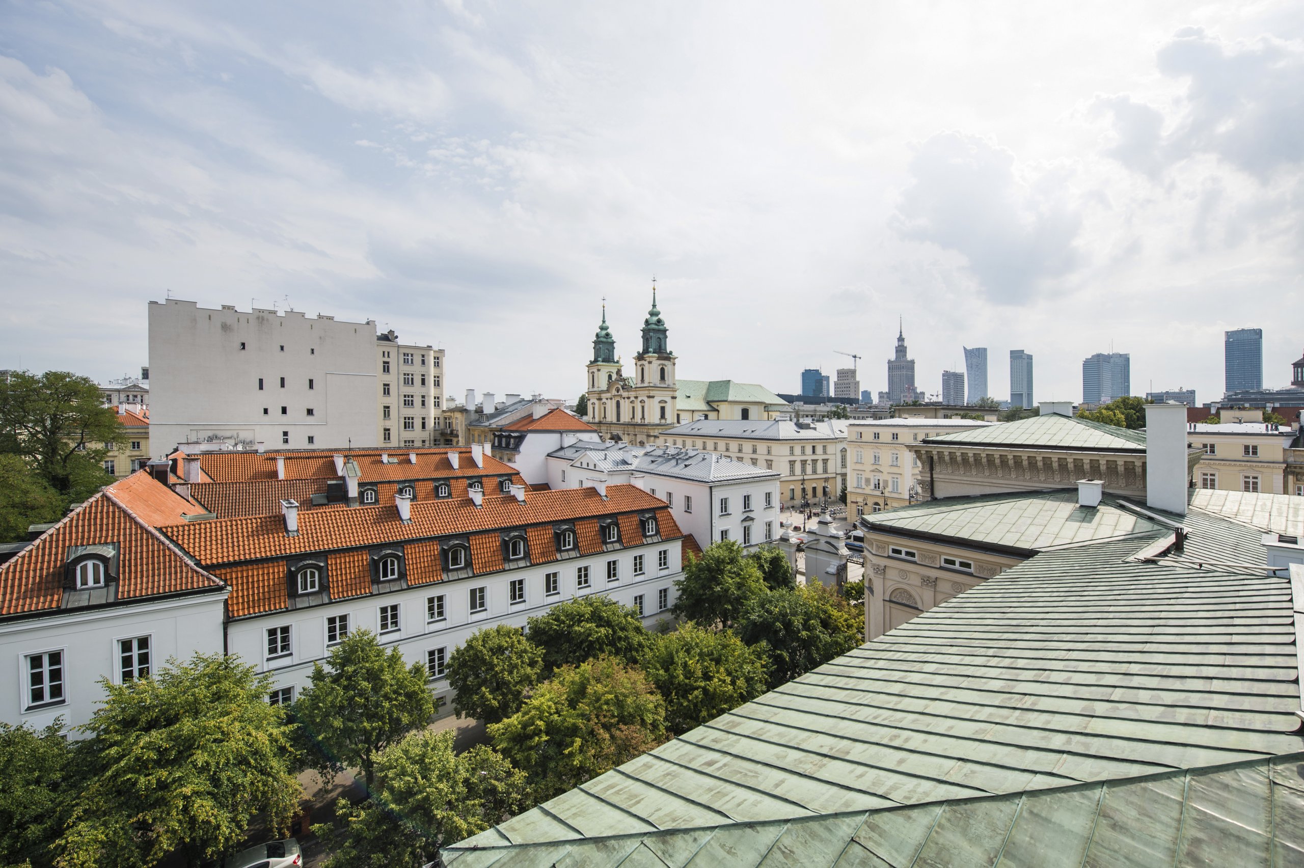 Zdjęcie przedstawia panoramę Warszawy widzianą ponad zielonym dachem budynku UW widocznym w prawym dolnym rogu kadru.