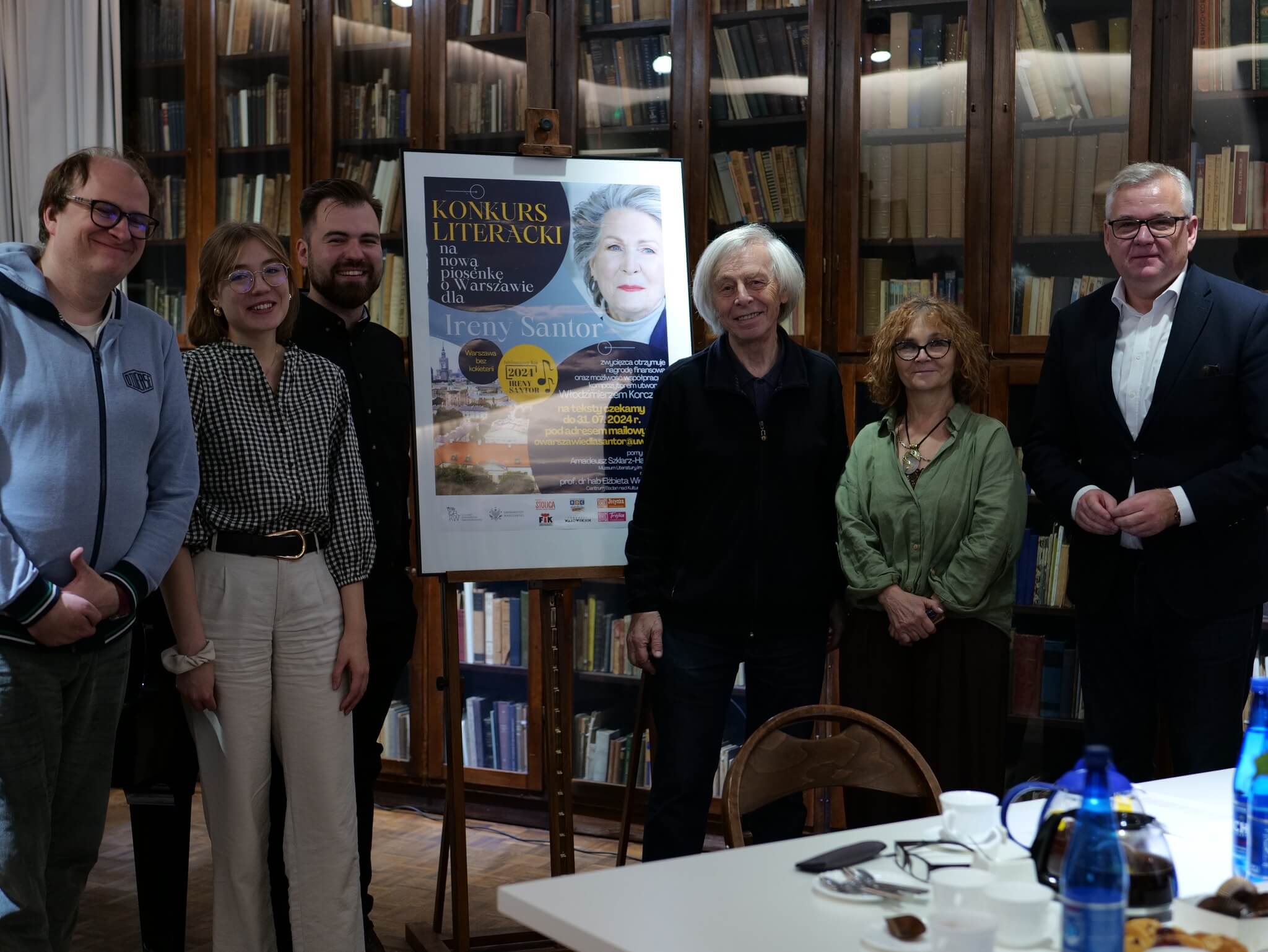 Jury konkursu na piosenkę o Warszawie dla Ireny Santor - na tle witryn z książkami we wnetrzach Muzeum Literatury stoją Od lewej: prof. Igor Piotrowski Igor Piotrowski, Olga Słomińska, Amadeusz Szklarz-Habrowski Amadeusz Maksymilian, Włodzimierz Korcz, prof. Elżbieta Wichrowska Elżbieta Wichrowska, Artur Andrus. W środku widoczny plakat konkuru umieszczony na sztaludze.