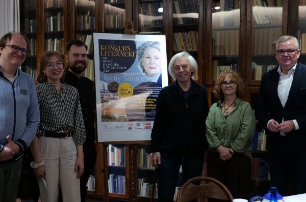 Jury konkursu na piosenkę o Warszawie dla Ireny Santor - na tle witryn z książkami we wnetrzach Muzeum Literatury stoją Od lewej: prof. Igor Piotrowski Igor Piotrowski, Olga Słomińska, Amadeusz Szklarz-Habrowski Amadeusz Maksymilian, Włodzimierz Korcz, prof. Elżbieta Wichrowska Elżbieta Wichrowska, Artur Andrus. W środku widoczny plakat konkuru umieszczony na sztaludze.