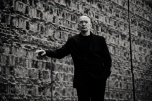 prof. Bogusław Stelmach. Zdjęcie monochromatyczne, mężczyzna w średnim wieku ubrany na czarno stoi oparty o szklaną balustradę na tle muru.