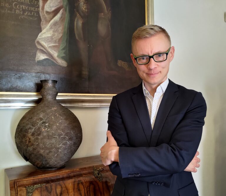 Marcin Dawidowicz. Mazowiecki Wojewódzki konserwator Zabytków. Mężczyzna w średnim wieku, w okularach stoi przy ścianie. W tle fragment obrazu olejnego.