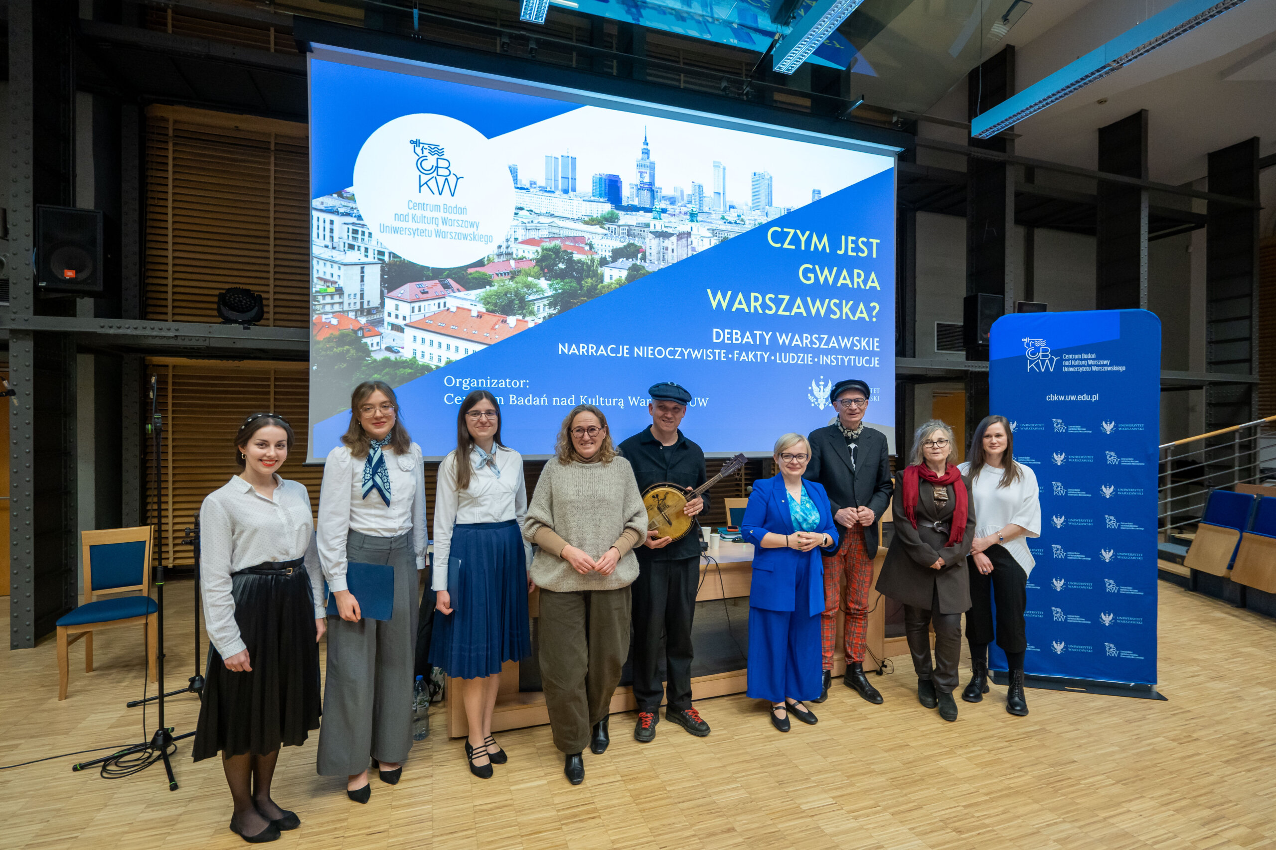 Debata warszawska: Czym jest gwara warszawska. Zdjęcie pamiątkowe: prelegenci Debaty i prowadzący wraz z chórzystkami Chóru Akademickiego UW oraz zespołem CBKW na tle plakatu Debaty.