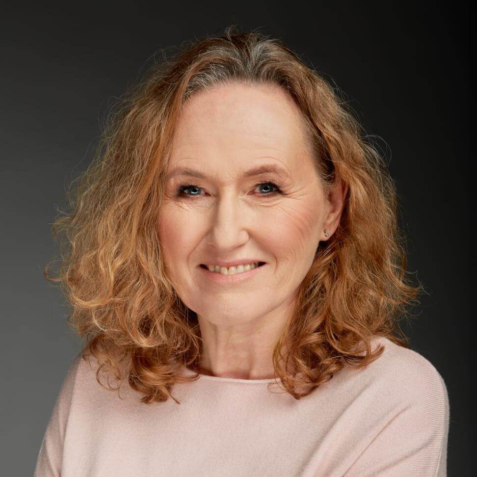 Ewa Malinowska-Grupińska. Portret uśmiechniętej kobiety w jasnej bluzce. Ma blond kręcone włosy do ramion.